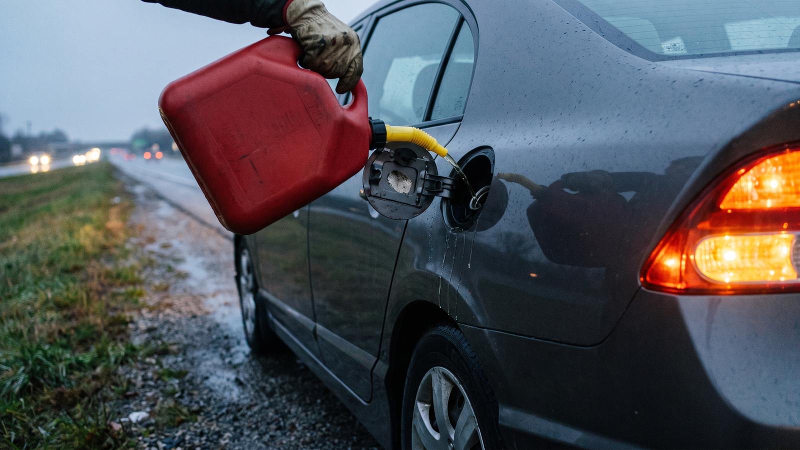 A gloved hand pouring fuel from a red gas can into a gray car's filler on a highway shoulder at dusk