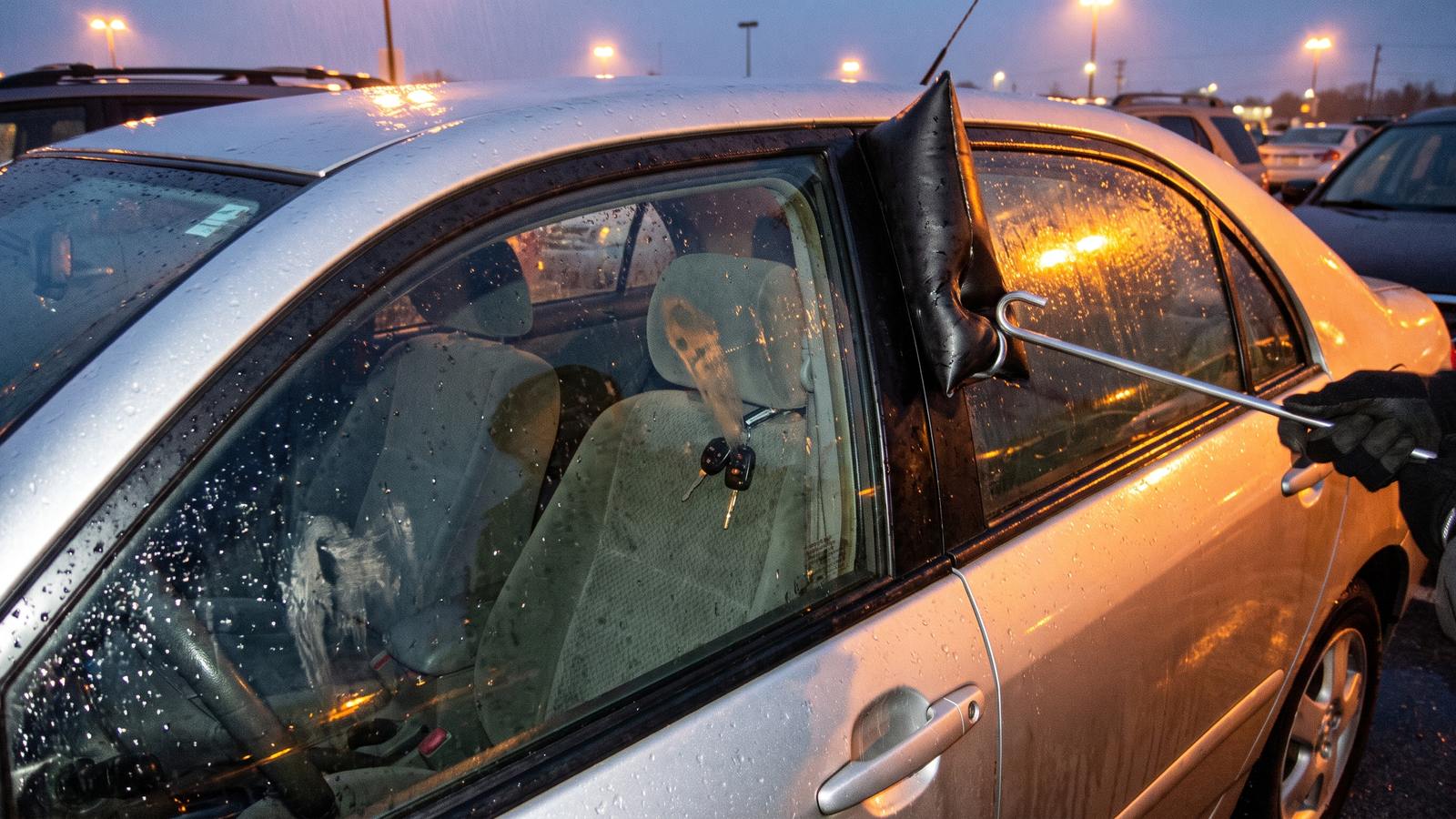 An air wedge and long-reach tool at the door frame of a locked silver sedan, with the keys visible inside